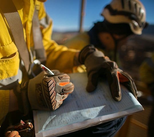 Worker wearing protective gloves and safety gear writing on a field form for wastewater and stormwater permitting compliance.